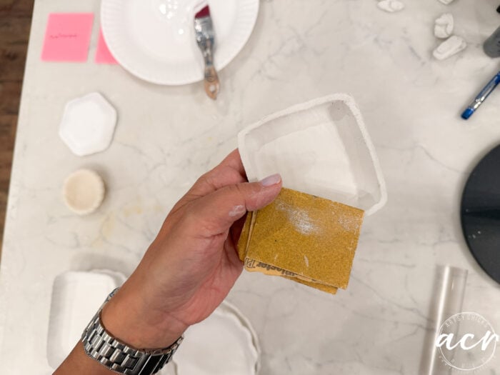 A hand holds sandpaper and a small ceramic dish, possibly made from Air Dry Clay. Art supplies, a paintbrush, and pink sticky notes are visible on a white marble table—perfect for making a comparison between clay brands while working on creative projects.