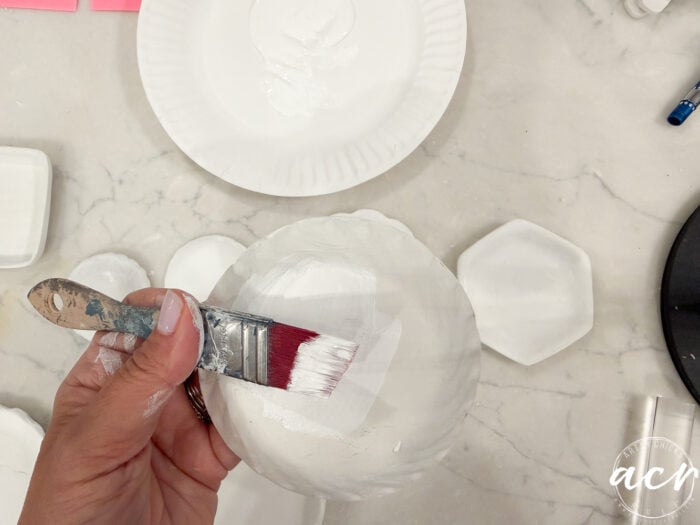 A hand holds a brush, painting a round object white—possibly made with one of the top air dry clay brands. Several white plates and painting supplies are on a marble surface in the background, perfect for anyone interested in air dry clay brands comparison.