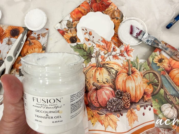 A hand holds a jar of Fusion decoupage gel near autumn-themed paper with pumpkins and leaves, surrounded by crafting tools, a brush, and a plate—perfect for an Air Dry Clay review or experimenting with different clay brands comparison in your seasonal crafts.
