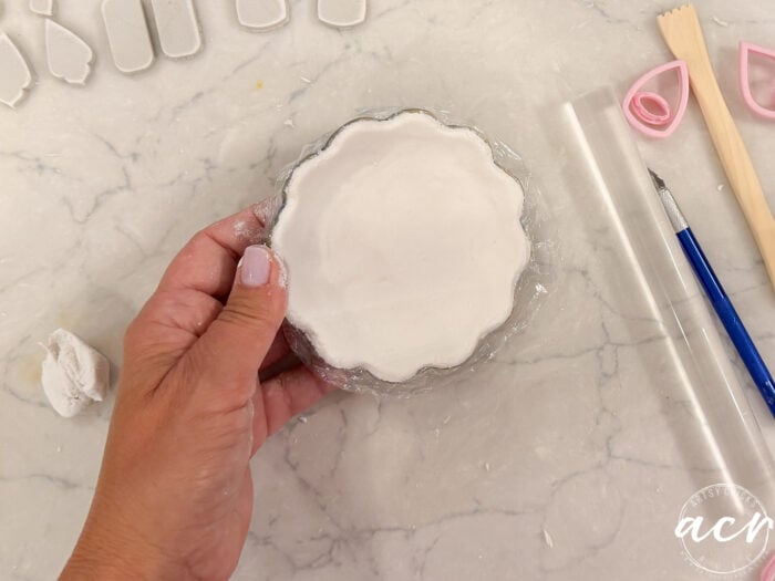 A hand holds a scalloped white clay dish made from air dry clay on plastic wrap. Clay tools and white clay pieces from different clay brands are on the marble surface, perfect for comparing brands and experimenting with textures.