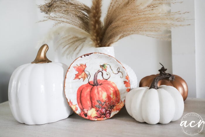 A fall-themed display with two white pumpkins, one bronze pumpkin, a pumpkin-decorated plate, and pampas grass in a white vase—all arranged on a light-colored surface—showcases the versatility of air dry clay for seasonal crafts.