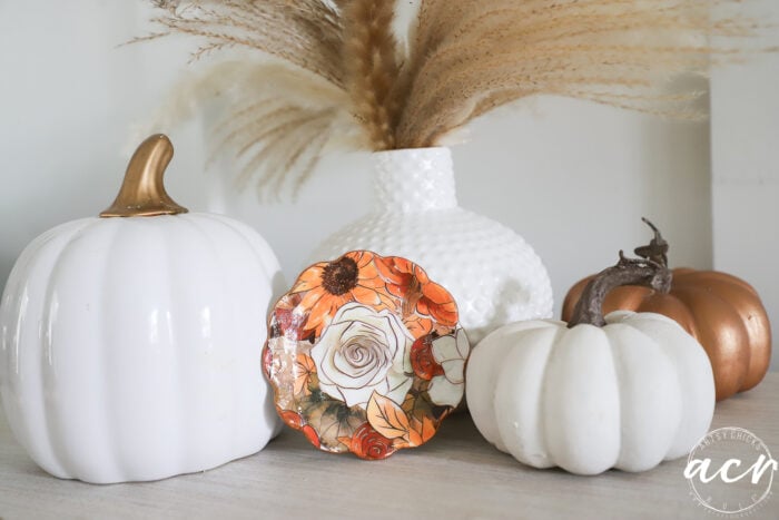 White and bronze pumpkins, a small floral plate, and a white vase with pampas grass are arranged on a light surface. Some of the decorative pumpkins appear to be crafted from air dry clay, adding an artisanal touch to the neutral and autumn colors. The image is styled in soft tones, with an acv watermark in the lower right corner.