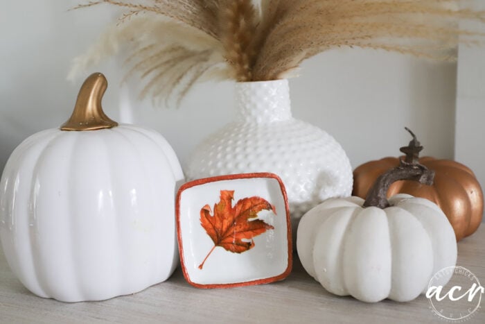 White and gold pumpkins, a small dish crafted from air dry clay with a painted orange leaf, and a white textured vase with pampas grass arranged together on a light surface for autumn decor.