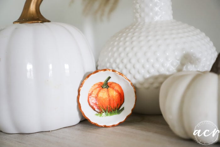 Three white decorative pumpkins and a white textured vase are arranged on a surface. In front, a small round dish with a painted orange pumpkin design is displayed. Perfect for an Air Dry Clay review or clay brands comparison, the pieces showcase subtle craftsmanship against a soft, neutral background.