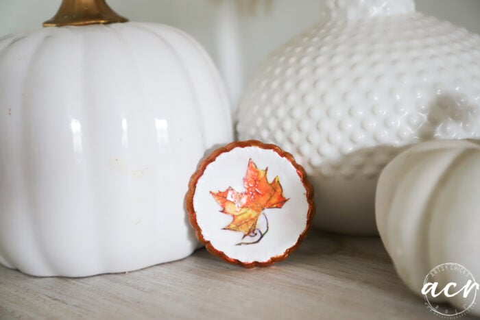 A small round dish sculpted from air dry clay with a painted autumn leaf sits on a wooden surface, surrounded by white ceramic pumpkins and vases, providing the perfect piece for any Air Dry Clay Review or Clay Brands Comparison.