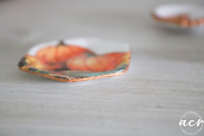 A small hexagonal dish with a pumpkin design, handcrafted from air dry clay, sits on a light surface; another dish is blurred in the background. The bottom right corner shows a circular watermark with the letters acn, highlighting this piece for those interested in Clay Reviews or comparing brands.