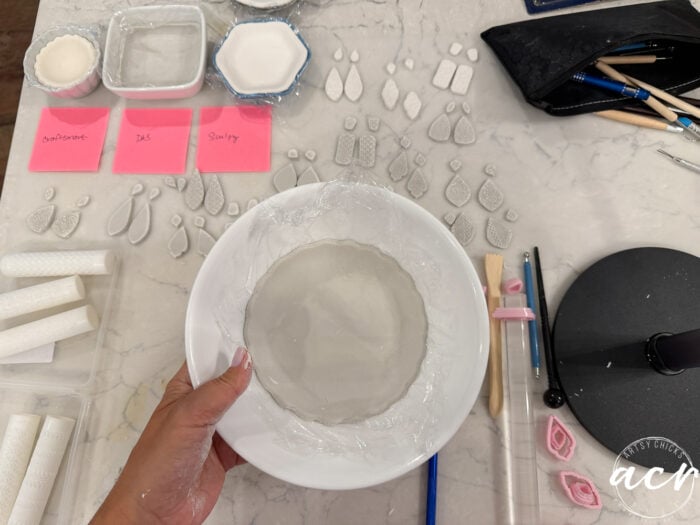 A hand holds a white plate with a round clay dish on it. Nearby are clay jewelry pieces, tools, and labeled pink sticky notes reading Bakehome, Daz, and Sculpy—showcasing an air dry clay comparison between popular air dry clay brands on a marble work surface.