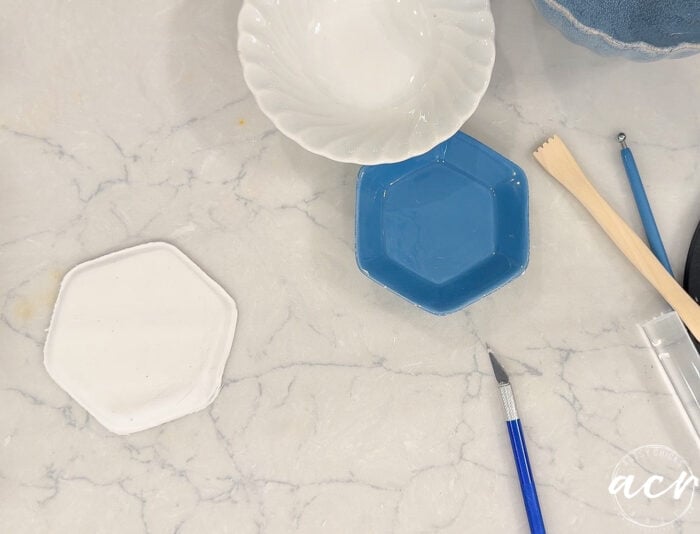 Hexagonal white and blue ceramic dishes on a marble surface, surrounded by sculpting tools, a paintbrush, and a white ceramic bowl—an inviting setup for those interested in air dry clay projects or seeking inspiration for their next clay brands comparison.