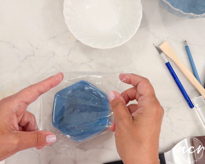 Hands cover a small blue hexagonal dish, crafted from air dry clay, with plastic wrap on a marble surface. Nearby are a white bowl, sculpting tools, and a blue fabric item—perfect for anyone interested in clay brands or seeking tips for brand comparison.
