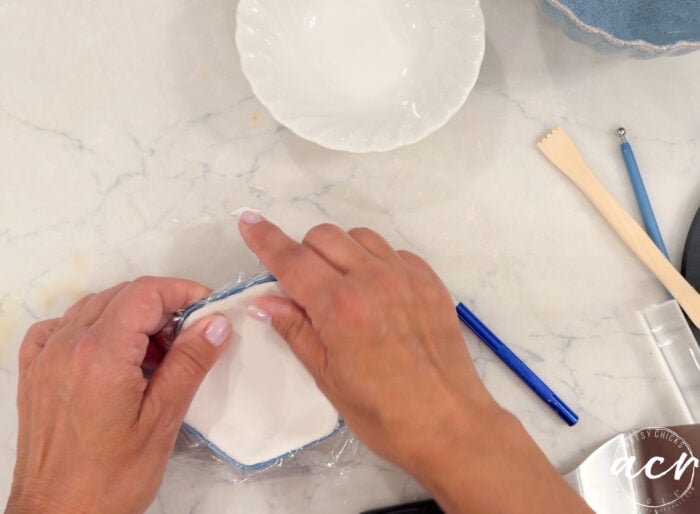 Two hands smooth plastic wrap over a small square dish of air dry clay on a marble countertop, with a white bowl, blue pen, and wooden utensils nearby.