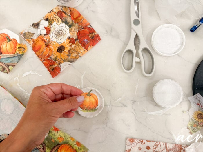 A hand holds a round object with a pumpkin image, sculpted using Air Dry Clay, surrounded by scissors, a glue container, and autumn-themed paper pieces on a white marble surface—a perfect scene for clay brands comparison and creative fall crafting.