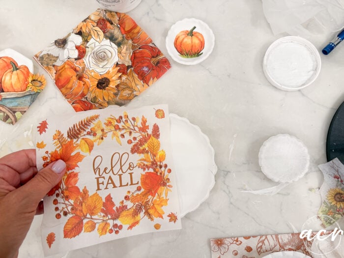 A hand holds a napkin with hello FALL and autumn leaves, surrounded by other fall-themed napkins, white bowls, and crafting supplies on a marble surface—an inviting scene perfect for comparing clay brands or incorporating air dry clay into your seasonal crafts.