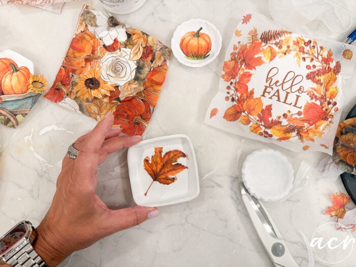 A hand holds a small dish with a fall leaf design, crafted from the best air dry clay. Autumn-themed napkins, one reading hello FALL, and craft supplies are scattered on a white surface.