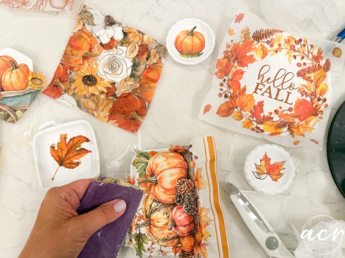 A hand sanding a pumpkin-decorated tile made with air dry clay on a table with autumn-themed napkins, scissors, and small plates, featuring fall leaves and pumpkins—perfect for those comparing brands of air dry clay for their seasonal crafts.