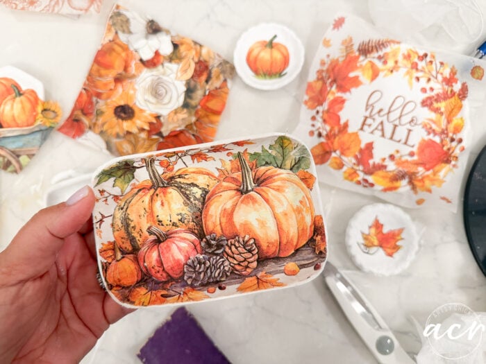 A hand holds a small tray decorated with pumpkins and pinecones. Autumn-themed napkins, scissors, and craft supplies—including air dry clay from various brands—are on a white table, reflecting a fall crafting project.