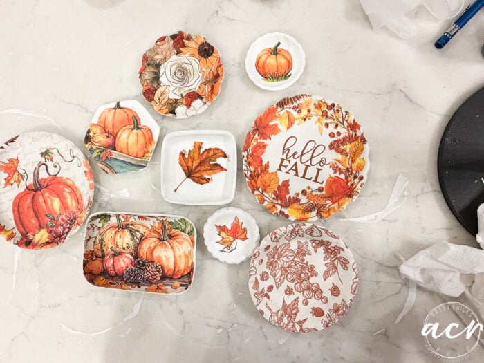 Assorted ceramic dishes with autumn-themed designs, such as pumpkins, leaves, and hello FALL text, are arranged on a white surface with some paper scraps and a partial logo in the bottom right corner. Perfect for those comparing brands or exploring different clay brands, these charming pieces showcase the versatility of air dry clay for seasonal crafting projects.