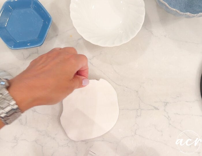 A hand wearing a silver watch holding a piece of white air dry clay on a marble surface, with blue and white dishes nearby—perfect for those interested in air dry clay or conducting a clay comparison between popular clay brands.