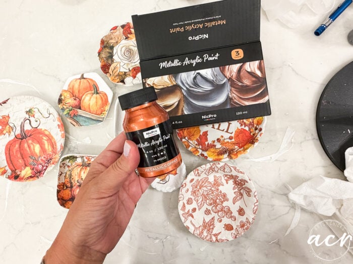 A hand holds a bottle of metallic copper acrylic paint over decorative fall-themed plates, with a box of metallic acrylic paints and painting supplies nearby—perfect for creative projects like comparing air dry clay brands or experimenting with various air dry clay techniques.