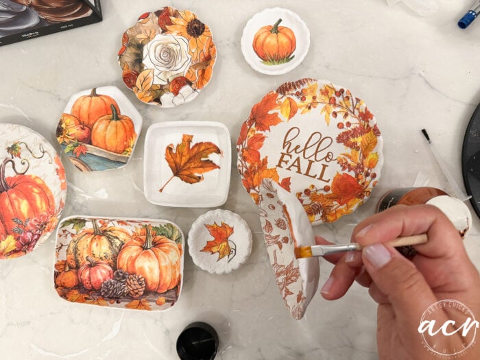 A hand paints glue on a dish decorated with autumn-themed napkins featuring pumpkins, leaves, and Hello Fall text, surrounded by other similarly decorated plates on a white surface—perfect for showcasing your latest Air Dry Clay creations or even making a comparison of different clay brands in seasonal craft projects.