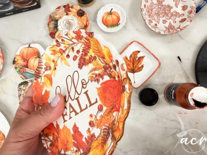A hand holds a pumpkin-shaped paper with Hello Fall and autumn leaves, surrounded by fall-themed plates and crafting supplies on a table—perfect inspiration for your next Air Dry Clay project or even a creative Clay Brands Comparison.