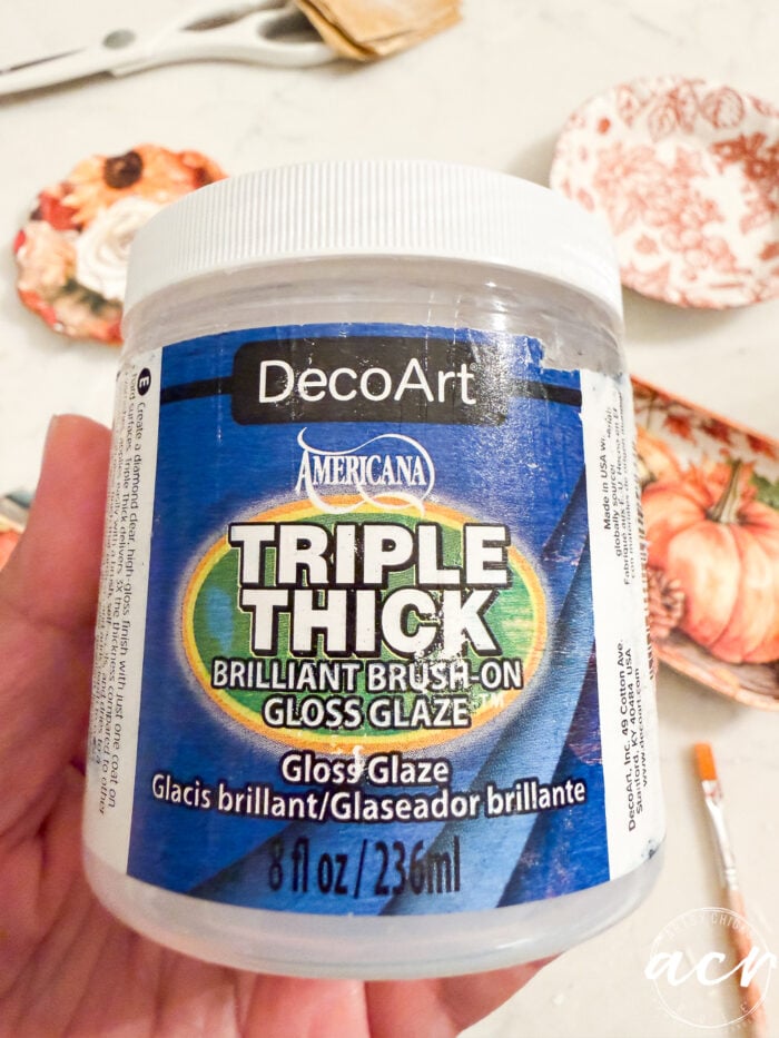 A hand holds a container of DecoArt Americana Triple Thick Brilliant Brush-On Gloss Glaze, 8 fl oz, with craft materials, air dry clay creations, and a paintbrush in the blurred background—perfect for comparing brands and achieving a flawless glossy finish.