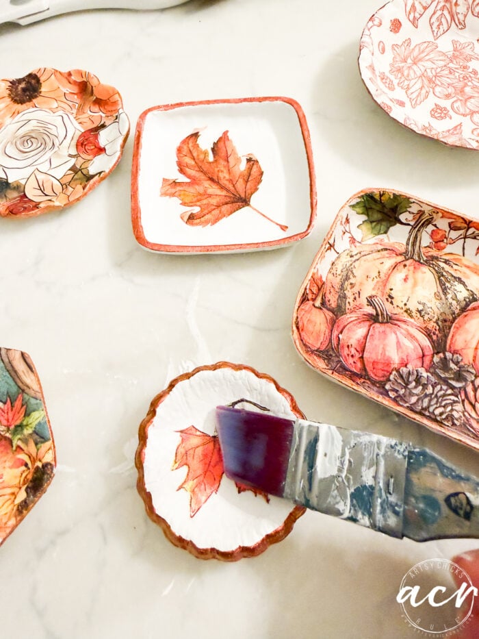 A hand uses a brush to paint glue over a small dish with a leaf design, showcasing the versatility of air dry clay. Other autumn-themed dishes with pumpkins and leaves, perfect for an air dry clay review, are arranged on a light surface.