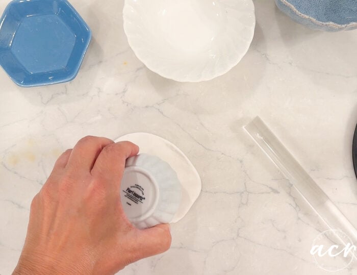 A hand presses a white bowl upside down onto white clay on a marble surface, with a blue dish, a clear tube, and another white bowl nearby—perfect for anyone interested in comparing Air Dry Clay brands.