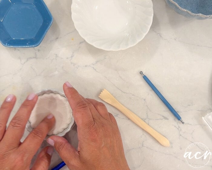 Two hands press white clay, such as a popular Air Dry Clay, into a small round dish on a marble surface, surrounded by sculpting tools and various empty dishes—an inviting scene for anyone interested in clay brands or eager for a brand comparison.