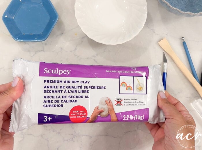 Hands hold a 2.2 lb package of Sculpey Air Dry Clay on a marble table, surrounded by clay tools, a white bowl, and a blue hexagonal dish. The package label is shown in English, French, and Spanish—a great choice if you’re looking to compare brands or write your next clay review.