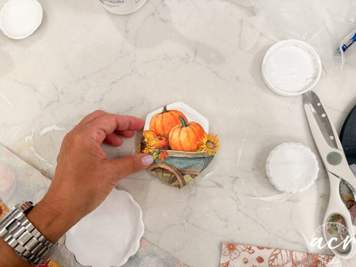 A hand holds a small decorative plate with painted pumpkins and sunflowers, showcasing a creative air dry clay project. Art supplies, a watch, scissors, and a palette are arranged on the white surface—perfect for anyone interested in experimenting with different clay brands or looking for an inspiring comparison of materials.