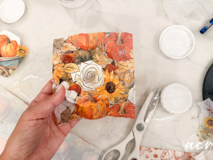 A hand holds a square piece of paper with an autumn-themed design featuring pumpkins, sunflowers, and a white rose; scissors, air dry clay, and other crafting supplies are on the table in the background—perfect for comparing brands or experimenting with different clay brands for your next seasonal project.
