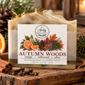 A bar of handcrafted natural soap labeled Autumn Woods sits on a wooden surface, surrounded by pinecones, orange slices, cinnamon sticks, and autumn leaves. The label features fall foliage, oranges, and spices—capturing the essence of all natural soaps inspired by the cozy scents of fall.