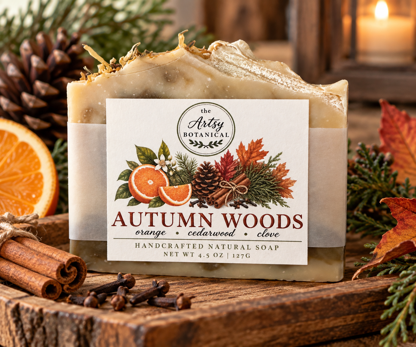 A bar of handcrafted natural soap labeled Autumn Woods sits on a wooden surface, surrounded by pinecones, orange slices, cinnamon sticks, and autumn leaves. The label features fall foliage, oranges, and spices—capturing the essence of all natural soaps inspired by the cozy scents of fall.