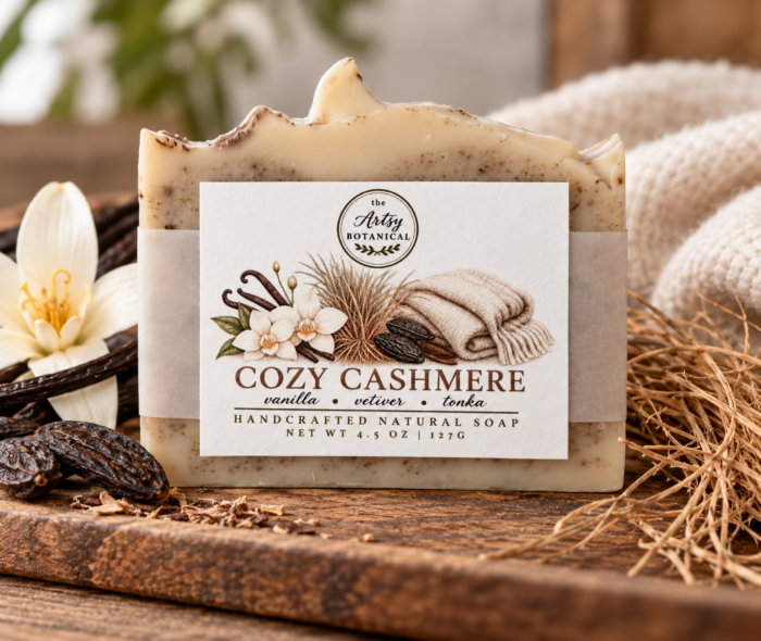 A handcrafted soap bar labeled Cozy Cashmere with images of vanilla, flowers, and a cozy blanket on a wooden surface, surrounded by natural elements and a flower. This Natural Soap captures the essence of comfort and warmth, perfect for those who appreciate all natural soaps inspired by the scents of fall.