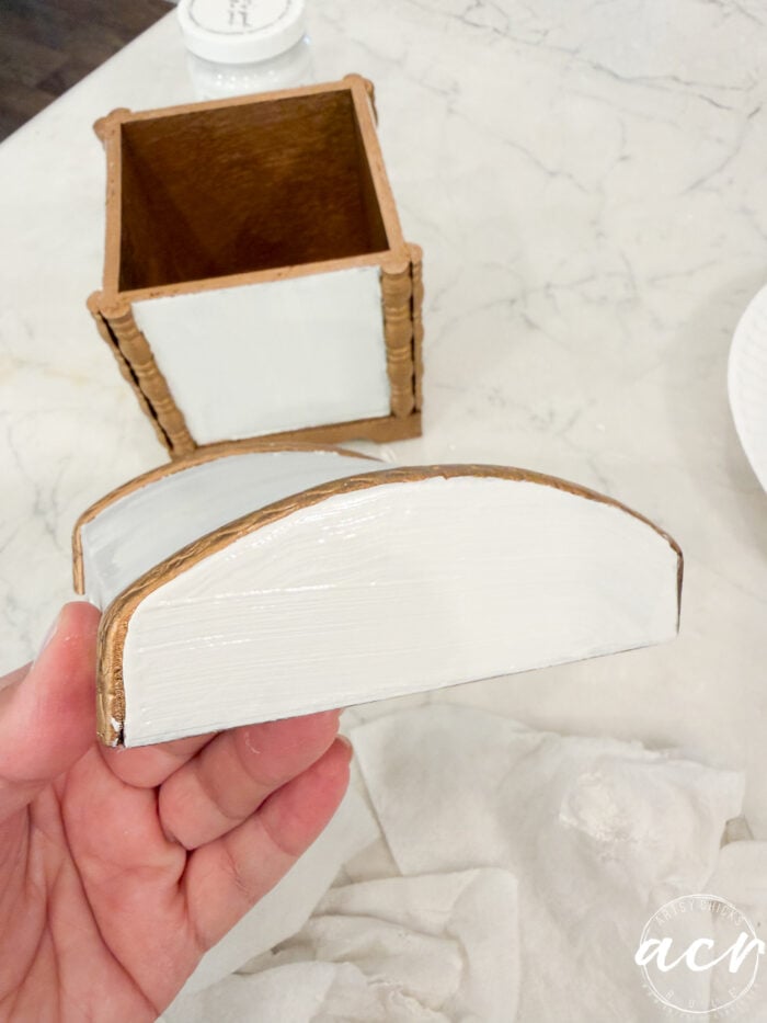 A hand holds a painted wood piece with white and gold edges, part of the Flipping Smalls #4 collection, near a matching rectangular container on a white surface with a paper towel and a round plate in the background.