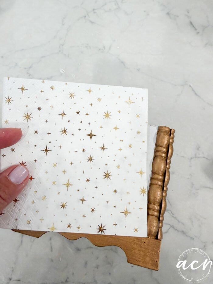 A hand holds a white napkin with gold star patterns, placed in a decorative gold napkin holder on a white marble surface—an elegant touch that would appeal to collectors of unique home décor and Flipping Smalls collectibles.