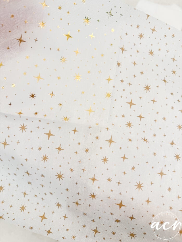White tissue paper with a pattern of small gold stars and dots scattered across the surface. The paper is slightly translucent and folded, featuring a subtle Flipping Smalls watermark logo in the bottom right corner.