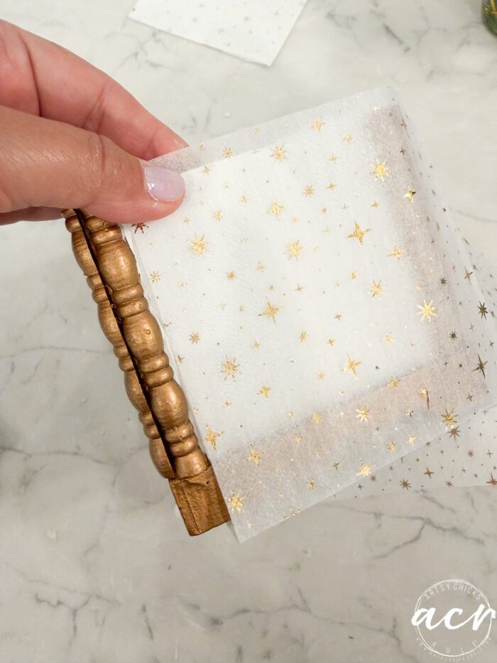 A hand holds a wooden object wrapped in translucent white paper with gold stars and dots—a charming find from Flipping Smalls. The background is a light-colored marble surface.