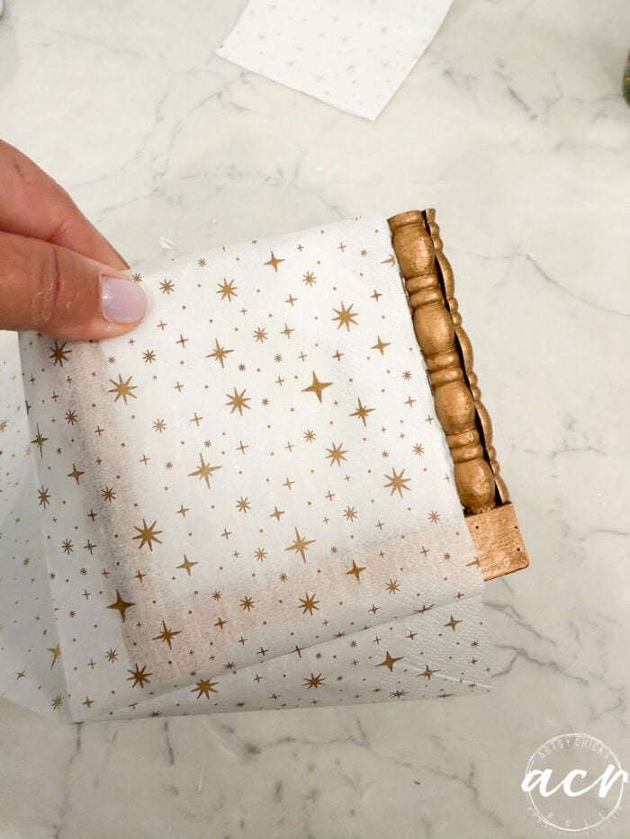A hand holds a festive napkin with gold stars, wrapping it around a small, decorative wooden frame on a marble surface—a charming scene perfect for Flipping Smalls enthusiasts seeking creative holiday décor ideas.