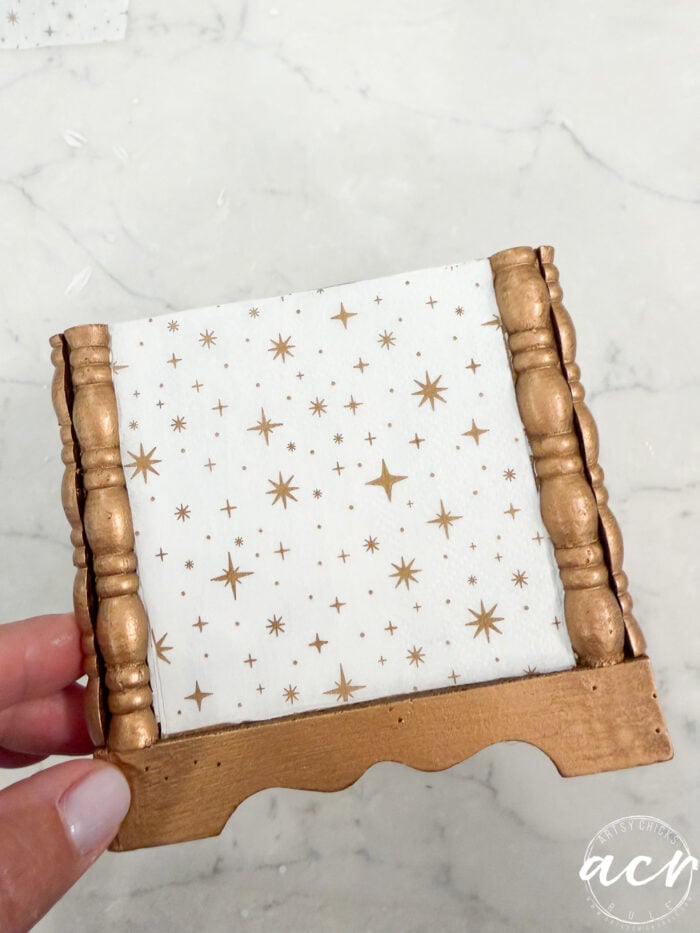 A hand holds a small decorative item with gold-painted edges, perfect for Flipping Smalls enthusiasts. The center features a white background with a gold star pattern, while the surface beneath is a light marble texture.