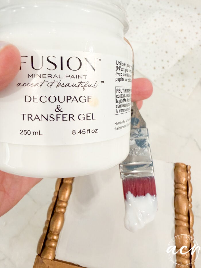 A hand holds a jar of Fusion Mineral Paint Decoupage & Transfer Gel from Flipping Smalls. A paintbrush with white gel is held nearby above a gold-edged surface.