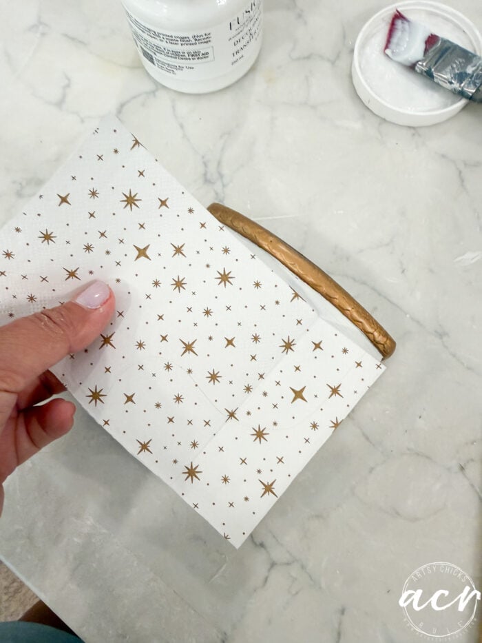 A hand holds a star-patterned napkin against a rectangular object with a gold handle on a marble surface; white glue, a brush, and a container lid are nearby—perfect for your next Flipping Smalls project.