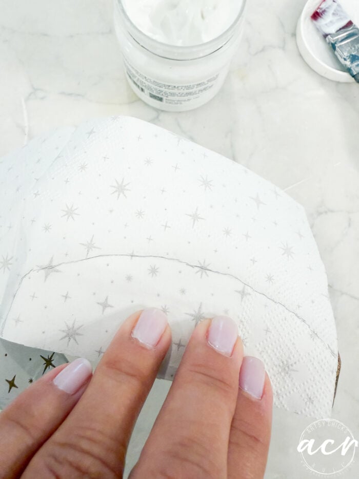 A hand with pale pink nails smooths a piece of white patterned paper onto a curved surface, channeling the creative spirit of Flipping Smalls, with a jar of white cream and art supplies in the background.