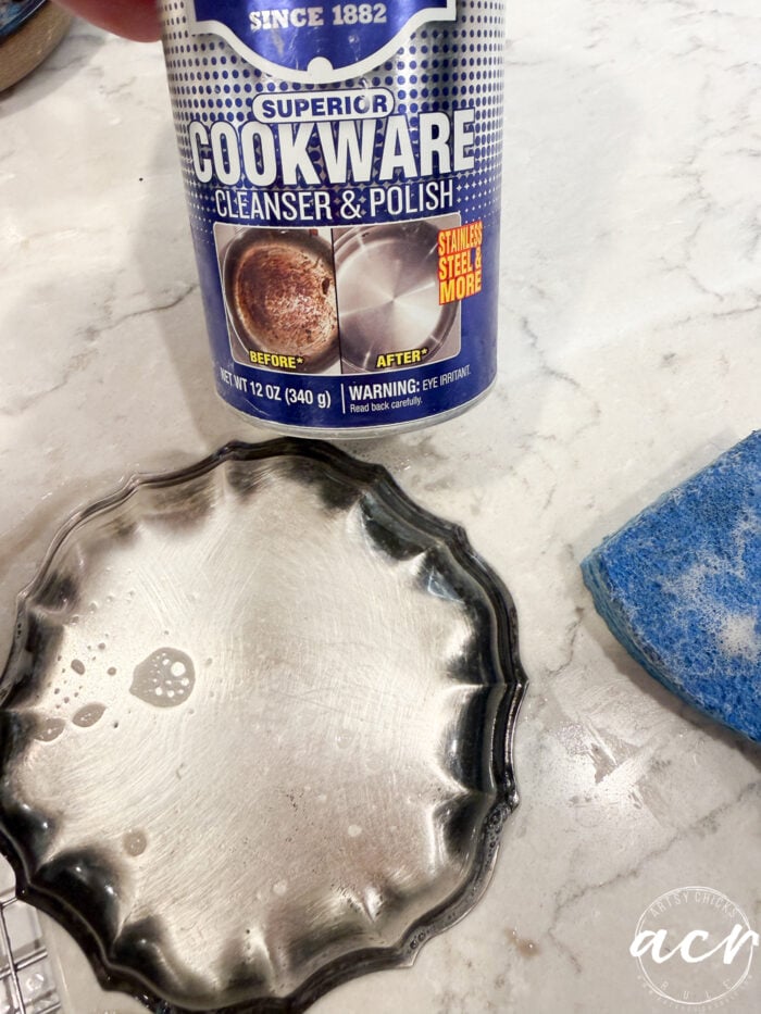 A can of Flipping Smalls cookware cleanser and polish sits on a countertop near a blue sponge and a soapy metal dish, suggesting cleaning in progress.