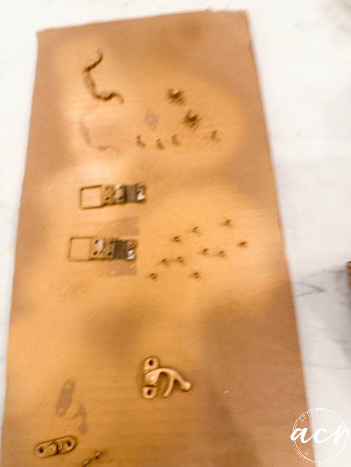Small metal hardware pieces, perfect for Flipping Smalls projects, lay on a brown, textured surface, partially sprayed with gold paint. The photo is slightly out of focus, giving a blurred effect.