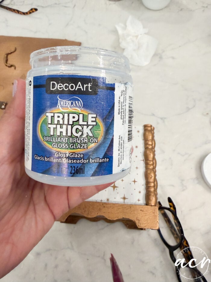 A hand holds a container of DecoArt Americana Triple Thick Gloss Glaze, 236ml, above a table with a small gold-painted shelf, a paintbrush, and a pair of glasses nearby—perfect supplies for your next Flipping Smalls project or to showcase on Flipping Smalls 4.
