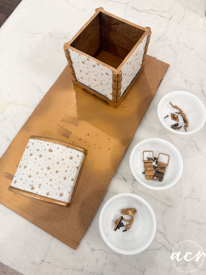 A decorated gingerbread house base with star patterns, a separate gingerbread roof, and three small white bowls holding various gingerbread and candy pieces sit on a white surface—an adorable scene perfect for Flipping Smalls #4 collectors or anyone seeking charming holiday collectibles.