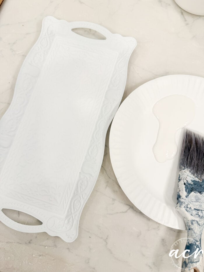 A white embossed tray sits on a marble surface next to a paper plate with white paint and a paintbrush with blue and white paint on its bristles, ready for another creative project inspired by Flipping Smalls.