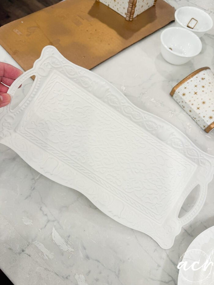 A hand holds a decorative white tray with ornate patterns, above a marble countertop—a perfect scene for Flipping Smalls enthusiasts. Nearby are white cups and star-patterned containers, adding charm to this stylish display.