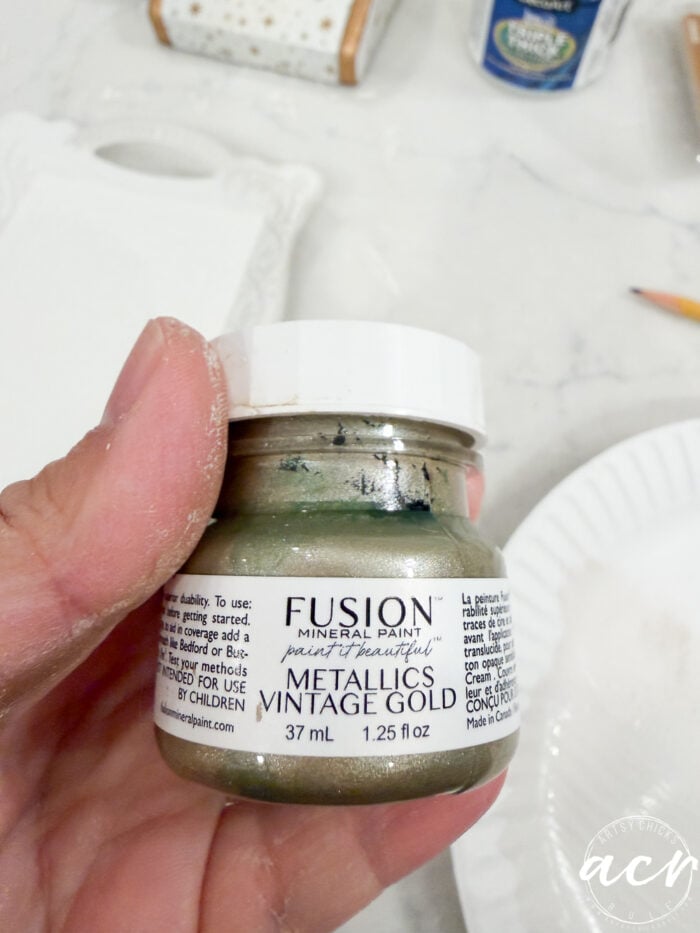 A hand holds a small 37 ml jar of Fusion Mineral Paint Metallics in Vintage Gold, with a white label and some paint residue on the lid—perfect for your next Flipping Smalls project.